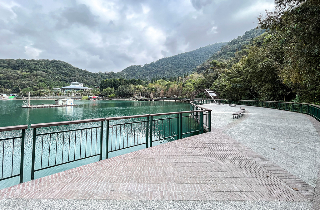 平坦且開闊的自行車步道，緊鄰日月潭。而佇立在不遠處的落羽松為畫面增添層次感。遠處的纜車站與連綿山巒相映，構成一幅自然與人文和諧交融的風景畫，讓人嚮往騎行其中，感受壯麗與寧靜。